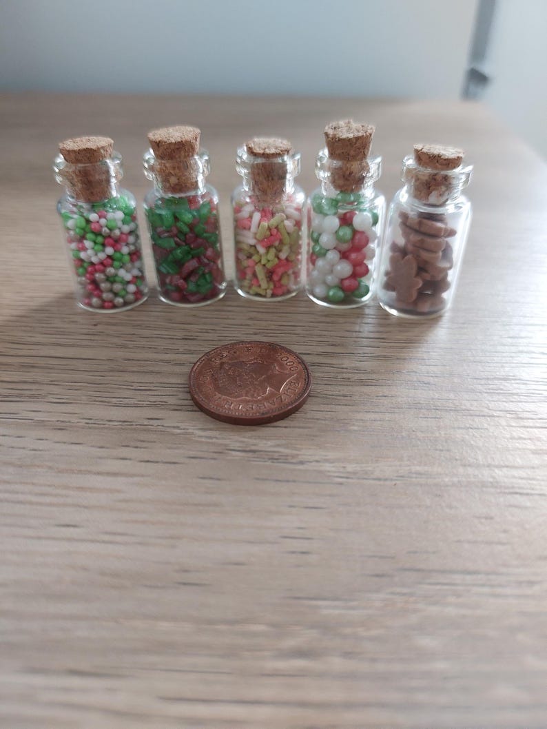 May include: Five small glass jars with cork lids filled with various colorful sprinkles and cookie shapes. The jars are lined up on a wooden surface with a copper coin in front of them.