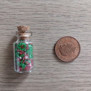 May include: A small glass bottle with a cork stopper filled with red and green sugar sprinkles. The bottle is next to a British 2000 penny coin.