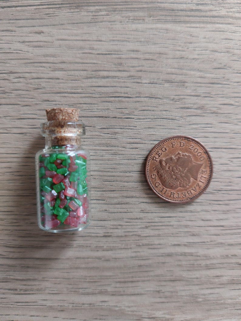 May include: A small glass bottle with a cork stopper filled with red and green sugar sprinkles. The bottle is next to a British 2000 penny coin.