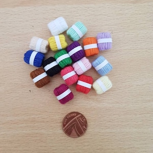 May include: A collection of 15 small balls of yarn in various colours, including white, light blue, yellow, green, purple, orange, red, brown, and black. The yarn balls are arranged in a circle on a wooden surface. A copper penny is in the centre of the circle for scale.