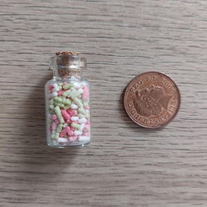 May include: A small glass jar filled with colorful sprinkles. The jar has a cork lid and is about the size of a British one pence coin.