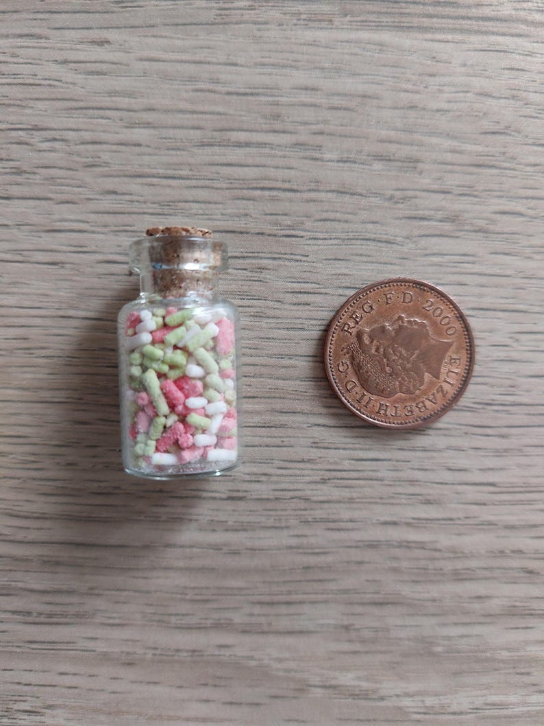 May include: A small glass jar filled with colorful sprinkles. The jar has a cork lid and is about the size of a British one pence coin.