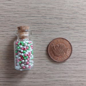 May include: A small glass bottle with a cork stopper filled with pink, green, and silver sprinkles. A British 1 pence coin is next to the bottle for scale.