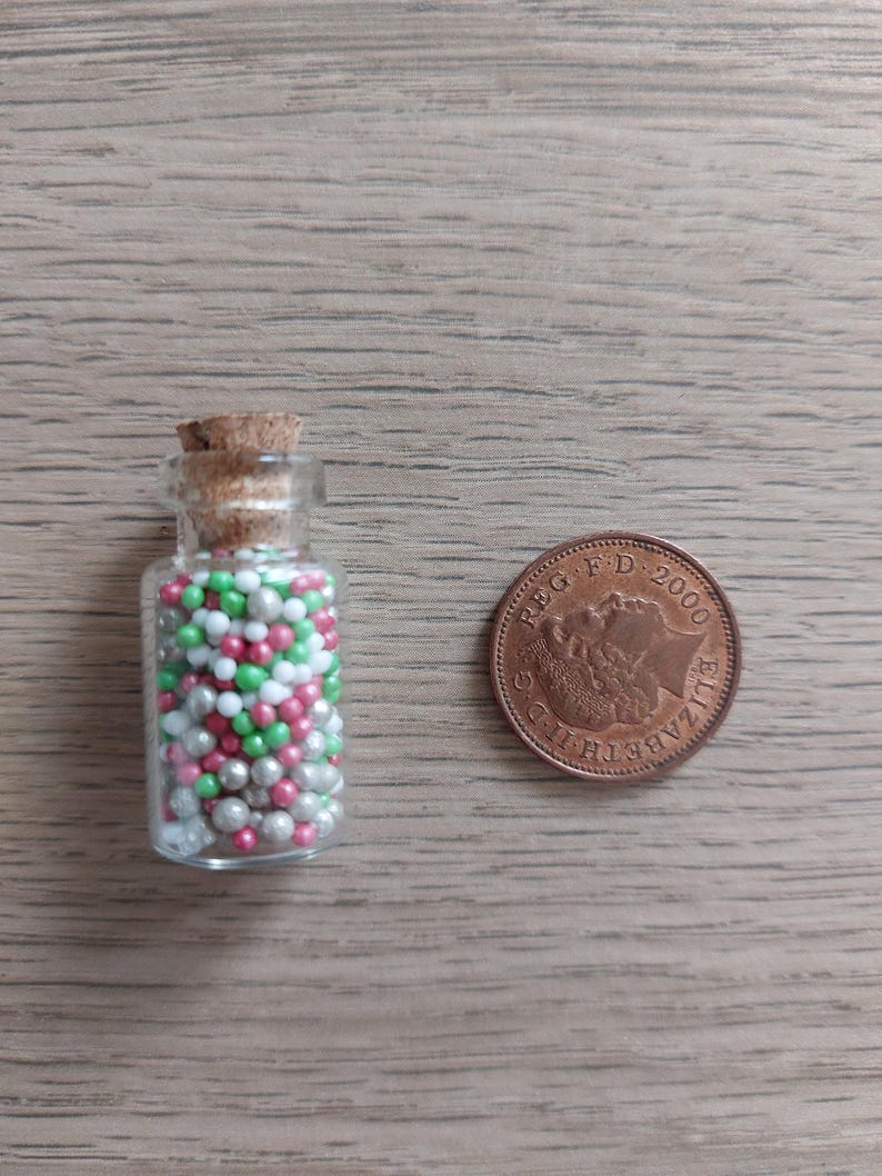 May include: A small glass bottle with a cork stopper filled with pink, green, and silver sprinkles. A British 1 pence coin is next to the bottle for scale.