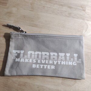 Könnte beinhalten: Eine graue Canvas-Reißverschlusstasche mit dem Aufdruck "FLOORBALL MAKES EVERYTHING BETTER" in Weiß.