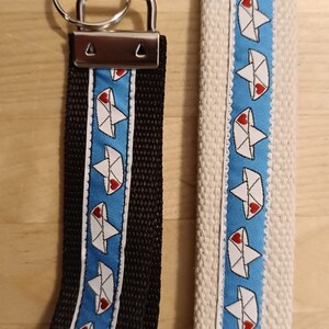 Könnte beinhalten: Zwei Schlüsselanhänger mit einem blau-weiß gestreiften Stoff mit einem weißen Papierboot-Design und einem roten Herzen. Die Boote haben einen weißen Umriss und ein rotes Herz in der Mitte. Die Schlüsselanhänger sind an silbernen Metallringen und Verschlüssen befestigt.