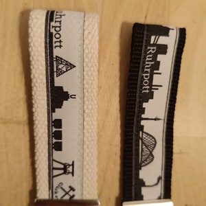 Könnte beinhalten: Zwei schwarz-weiße Schlüsselanhänger mit silbernen Schlüsselringen. Die Schlüsselanhänger haben einen weißen Hintergrund mit einer schwarzen Silhouette einer Stadtkulisse und dem Text "Ruhrpott" in Schwarz.