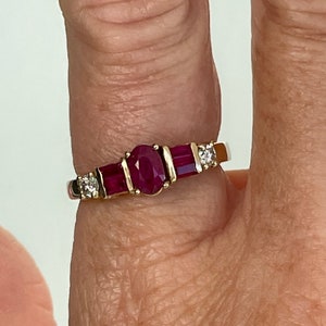 May include: A gold ring with a central oval ruby, two rectangular rubies, and two small diamonds. The ring's gold band highlights the red rubies and the diamonds' sparkle. The ring is a classic design.