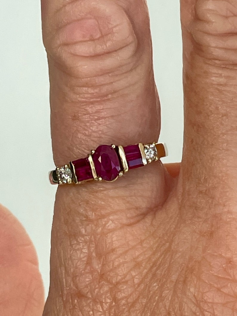 May include: A gold ring with a central oval ruby, two rectangular rubies, and two small diamonds. The ring's gold band highlights the red rubies and the diamonds' sparkle. The ring is a classic design.