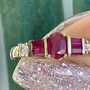 May include: A gold ring with an oval ruby centre stone, two rectangular rubies, and small diamonds. The ring has a classic design with a polished gold band and is set with multiple gemstones.