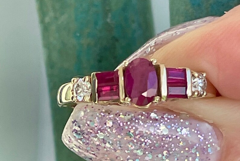 May include: A gold ring with an oval ruby centre stone, two rectangular rubies, and small diamonds. The ring has a classic design with a polished gold band and is set with multiple gemstones.