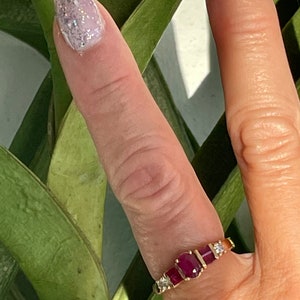 May include: A gold ring with three oval, deep red gemstones and smaller clear stones. The ring is set against green leaves and a light background. The ring is on a finger with glittery nail polish.