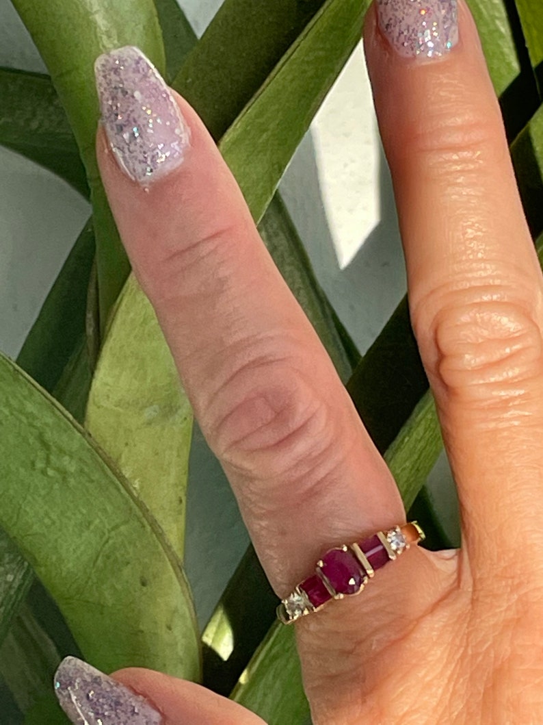 May include: A gold ring with three oval, deep red gemstones and smaller clear stones. The ring is set against green leaves and a light background. The ring is on a finger with glittery nail polish.