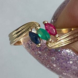 Può includere: Un anello d'oro con tre pietre preziose: uno smeraldo verde, uno zaffiro blu e un rubino rosso. L'anello ha un design ondulato.
