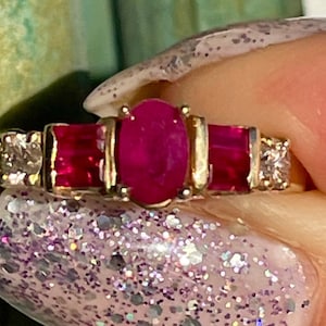 Può includere: Un anello d'oro con un rubino ovale centrale, affiancato da due rubini rettangolari e piccoli accenti di diamanti. Il design dell'anello è classico, con una finitura lucida e brillante.