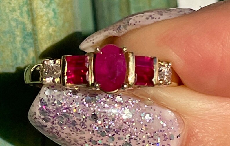 May include: A gold ring with a central oval ruby, two rectangular rubies, and diamond accents. The ring has a classic design with a polished finish. The rubies and diamonds are set in a gold band.