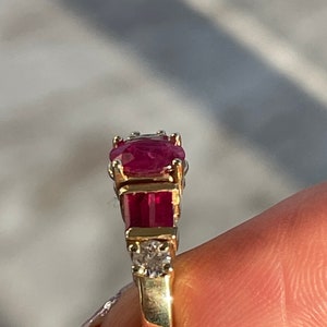 May include: A gold ring with a large oval ruby centre stone, two rectangular rubies, and small diamonds. The ring has a classic design with a polished gold band, showcasing the vibrant red gemstones.