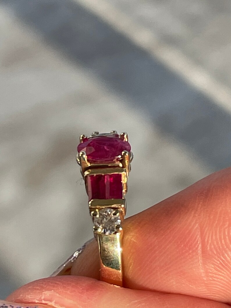May include: A gold ring with a large oval ruby centre stone, two rectangular rubies, and small diamonds. The ring has a classic design with a polished gold band, showcasing the vibrant red gemstones.
