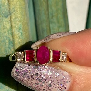 May include: A gold ring with a central oval ruby, two rectangular rubies, and two small diamonds. The ring's band is polished gold. The rubies are a deep red, and the diamonds sparkle. A classic piece of jewellery.