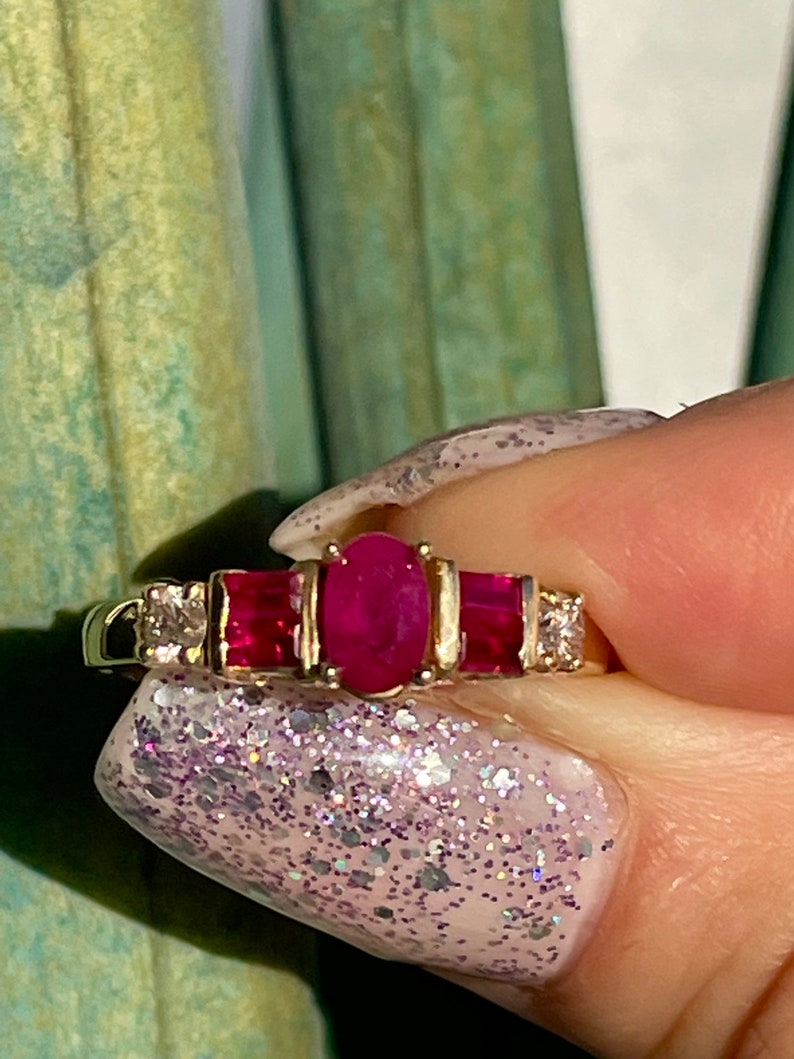 May include: A gold ring with a central oval ruby, two rectangular rubies, and two small diamonds. The ring's band is polished gold. The rubies are a deep red, and the diamonds sparkle. A classic piece of jewellery.