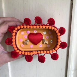 May include: A decorative, rectangular trinket dish with an orange base, adorned with red pom-poms and a heart-shaped accent. The dish features small dog figurines, flower-shaped sequins, and a red satin heart in the center.