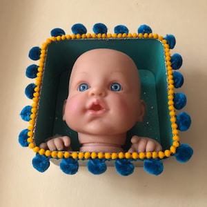 May include: A doll's head with blue eyes and an open mouth is set within a teal box. The box is trimmed with yellow pom-poms and a silver glitter border. The doll's hands are visible, resting on the box's edge.