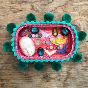 May include: A rectangular red tray decorated with dark green pom-poms and glitter. The interior features a white doll face, a yellow cherub, blue bird, Mona Lisa cameo, teal gems, and letter beads spelling out the phrase TAKE NO SHIT.