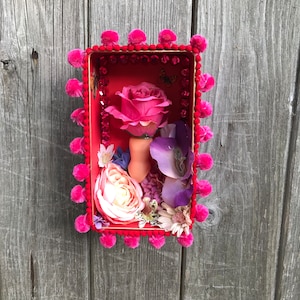 Peut inclure: Un arrangement floral rose, rouge et violet dans un cadre rouge avec une bordure en pompons roses. Les fleurs sont disposées dans une boîte avec un fond rouge et une surface scintillante.