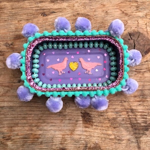 May include: A rectangular decorative tray with a purple interior featuring pink doves and heart accents. The tray is trimmed with teal beads, glitter, and lavender pom-poms. The tray is on a wooden surface.