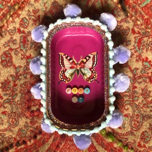 May include: A rectangular, magenta-colored trinket tray with a butterfly graphic and the words "FUCK IT" spelled out in colorful beads. The tray is trimmed with glitter, light blue pom-poms, and a light blue border.