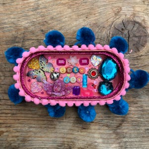 Puede incluir: Una bandeja ovalada con interior rosa y borde rosa brillante, decorada con pompones azules. La bandeja contiene varios abalorios, incluyendo una mariposa, un querubín, gemas azules y cuentas con letras que forman la frase SEEK JOY.