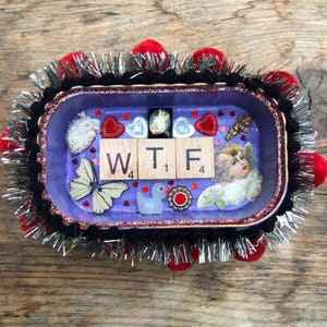 Puede incluir: Una bandeja rectangular morada decorada con un collage de una mariposa, un querubín, un corazón, un pato y tres fichas de madera con las letras WTF. El borde está adornado con pompones negros, espumillón plateado y pompones rojos.