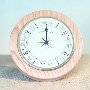 Può includere: Un orologio delle maree rotondo con cornice in legno chiaro. Il quadrante è bianco con numeri e testo neri che indicano i livelli delle maree. Al centro c'è una rosa dei venti. L'orologio misura circa 13 cm di diametro.