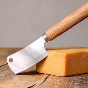 May include: A stainless steel cheese cleaver with a wooden handle rests on a block of cheddar cheese. The cheese is a vibrant yellow-orange color, and the cleaver is positioned at an angle, suggesting it has been used to cut the cheese. The background is a wooden surface.
