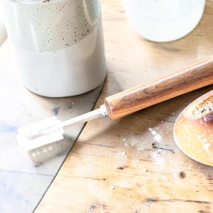 Puede incluir: Un cortador de masa de madera con una cuchilla y un mango de color claro descansa sobre una superficie de madera. También se ve un pan parcialmente rebanado y un recipiente de cerámica. La escena sugiere hornear o preparar alimentos.