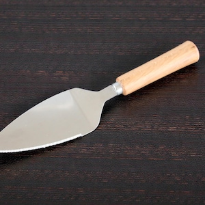 May include: A stainless steel cake server with a serrated edge and a light-colored wooden handle. The server is angled on a dark wood surface.
