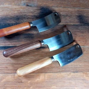 Puede incluir: Tres cuchillos de carnicero con mangos de madera sobre una superficie de madera. Las hojas de acero inoxidable tienen un agujero cerca de la parte superior. Los mangos son de maderas claras y oscuras. Para la cocina.