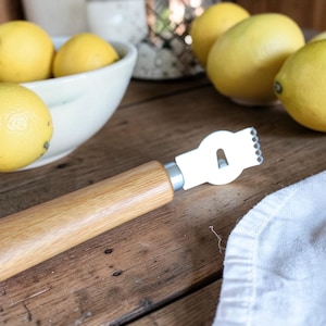 Könnte beinhalten: Ein Zitronenreißer mit Holzgriff und weißer Metallklinge, neben einer Schüssel mit Zitronen und losen Zitronen. Die Zitronen sind gelb und liegen auf einer Holzoberfläche. Ein weißes Tuch ist im Hintergrund.