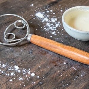 May include: A stainless steel wire whisk with a wooden handle sits on a dark wooden surface, beside a small ceramic bowl filled with a creamy substance. Flour is sprinkled around the whisk and bowl.