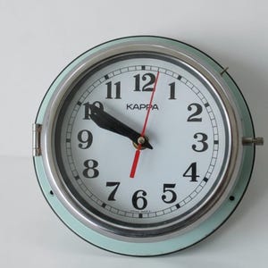 Könnte beinhalten: Eine runde Wanduhr mit weißem Zifferblatt, schwarzen Ziffern und schwarzen Zeigern. Die Uhr hat einen roten Sekundenzeiger und eine hellblaue Lünette. Das Wort "KAPPA" ist über der Mitte des Zifferblatts aufgedruckt.