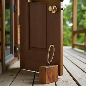 May include: A wooden doorstop with a rope handle is positioned to hold open a brown wooden door. The doorstop is a square block of wood with a natural finish. The door is on a wooden porch.