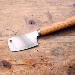 Könnte beinhalten: Ein Edelstahl-Hackmesser mit Holzgriff. Die Klinge ist glänzend und hat ein Loch in der Nähe der Oberseite. Der Griff ist aus hellem Holz. Das Hackmesser liegt auf einer Holzoberfläche.