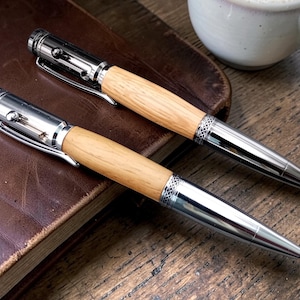 May include: Two stylish pens with light wood barrels and silver detailing are placed on a brown leather notebook. The pens feature a silver clip and tip. A white ceramic mug is visible in the background.