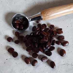 May include: A stainless steel portafilter with a wooden handle, filled with dark roasted coffee beans, sits next to a pile of loose beans. The beans are a rich brown color, scattered on a gray concrete surface.