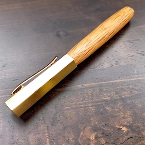 Può includere: Una penna in legno con cappuccio e clip in metallo color oro. Il fusto della penna presenta un motivo a venature del legno marrone chiaro. La penna è appoggiata su una superficie di legno marrone scuro.