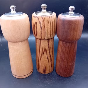 Puede incluir: Tres molinillos de pimienta de madera con tapas de acero inoxidable. Los molinillos varían en veta y color de la madera, desde marrón claro a oscuro. El diseño presenta una parte superior redondeada y un cuerpo cónico.