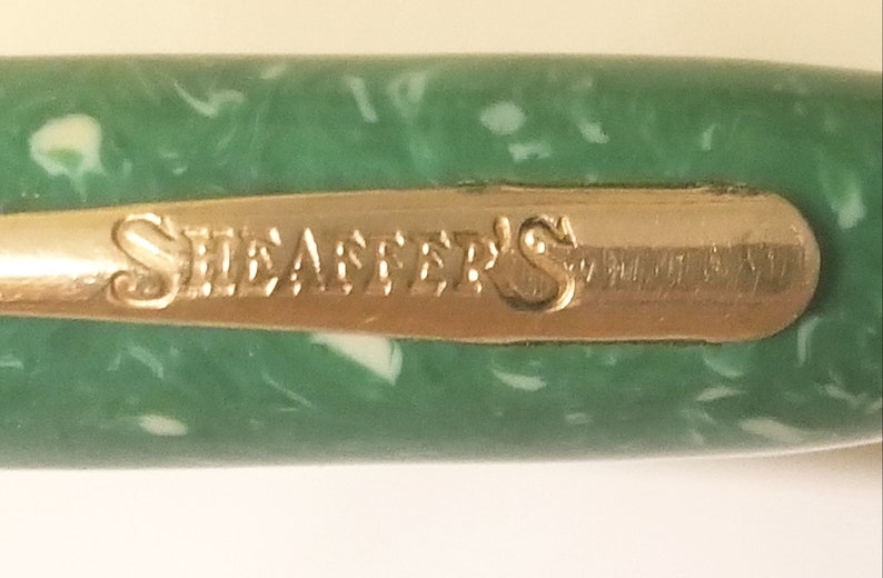 May include: Close-up of a green pen with a gold band that reads "Sheaffer's".