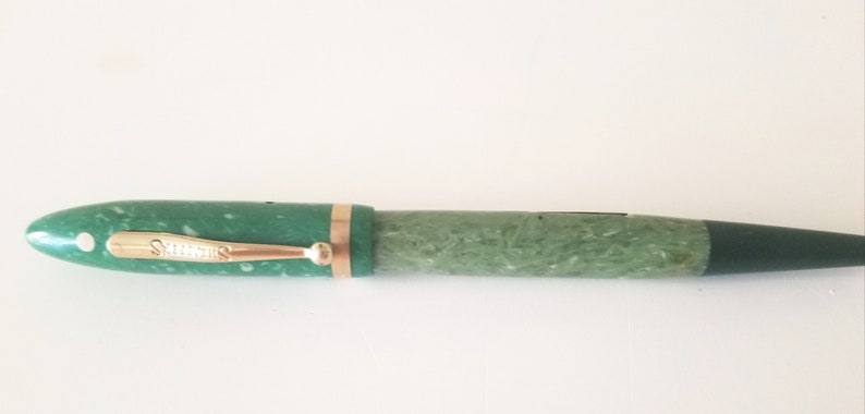 May include: A green and gold fountain pen with a marbled pattern. The pen has a gold trim and a black cap.