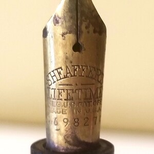 May include: Close-up of a gold-toned fountain pen nib with the inscription "Sheaffer's Lifetime Reg. U.S. Pat. Off. Made in U.S.A. 169827".