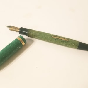 May include: A green and black fountain pen with a gold band. The pen is closed and the cap is off. The pen is on a white surface.